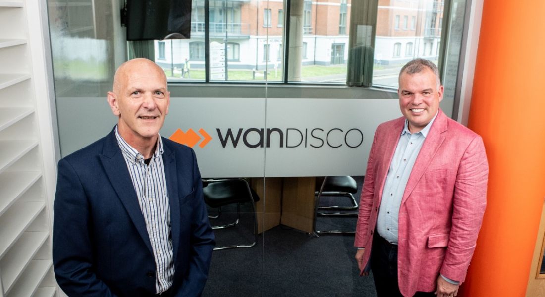 Two men stand on either side of athe WANdisco logo on a glass wall.