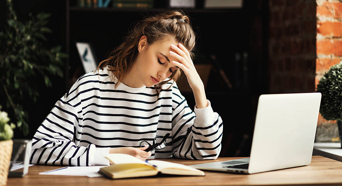 A young woman sitting at a desk holding her head, looking very tired, symbolising the effects of Covid-19 and long Covid.