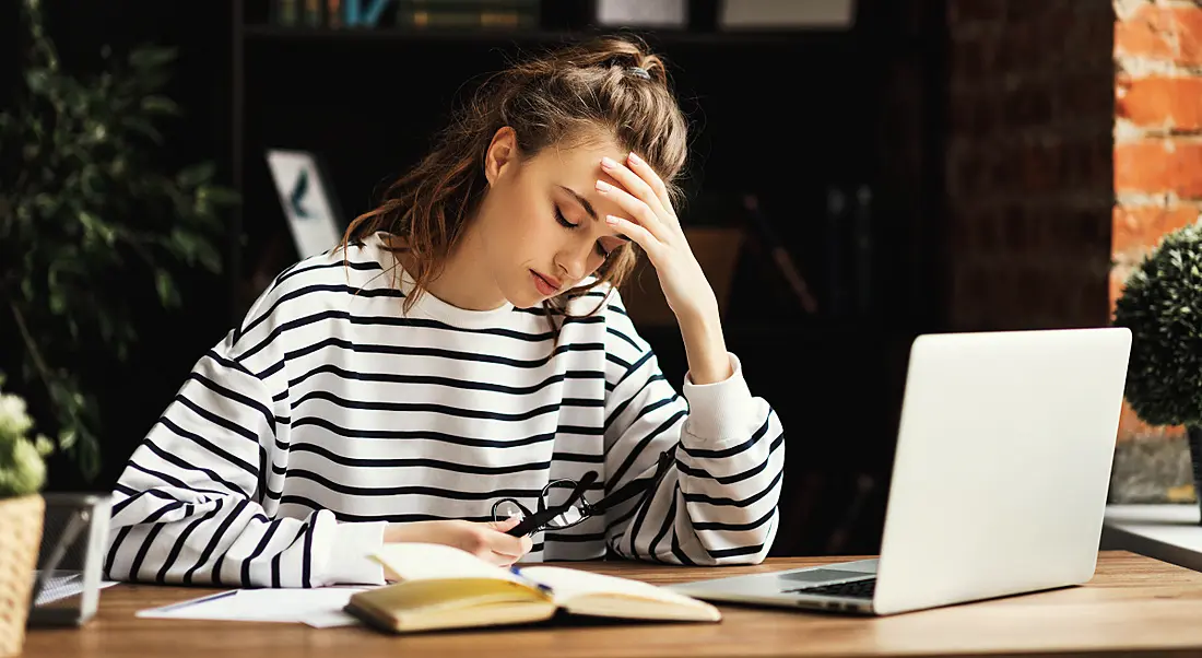 A young woman sitting at a desk holding her head, looking very tired, symbolising the effects of Covid-19 and long Covid.