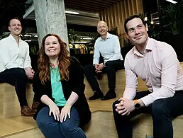 Photograph of three men and one woman standing together smiling for the camera, all wearing business outfits. They are Stephen McLoughlin, Paul James-Martin, Madeline Rogers and Chris Reid.