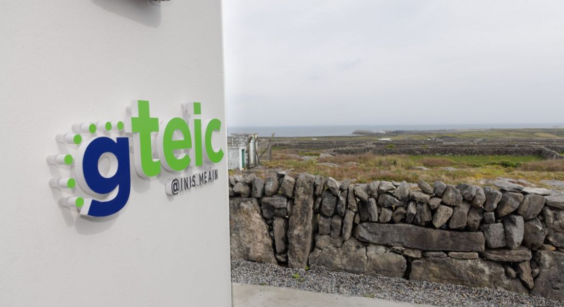 A sign on a wall says Gteic @ Inis Meáin, in front of a scenic rural background.