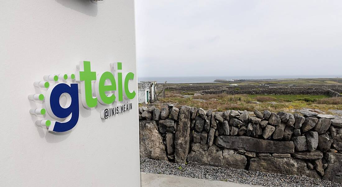 A sign on a wall says Gteic @ Inis Meáin, in front of a scenic rural background.