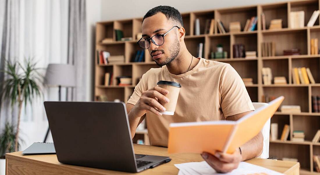 A young man holding an orange booklet while looking at a laptop and holding a coffee. There is a large bookshelf behind him.