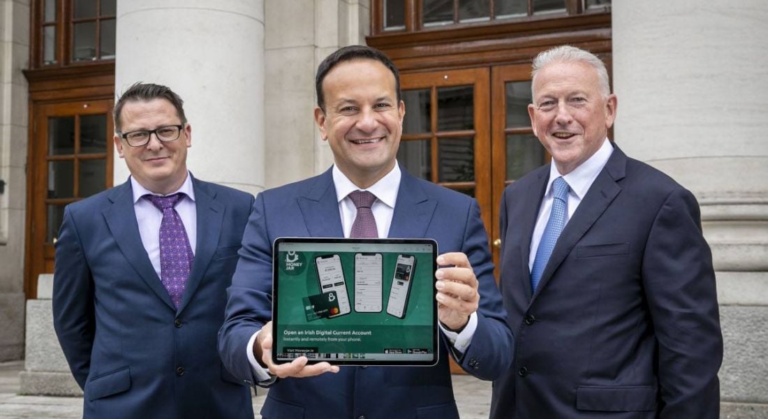 Three men stand outside Government buildings. In the centre is Leo Varadkar, holding up a tablet with the Money Jar website open on the screen.