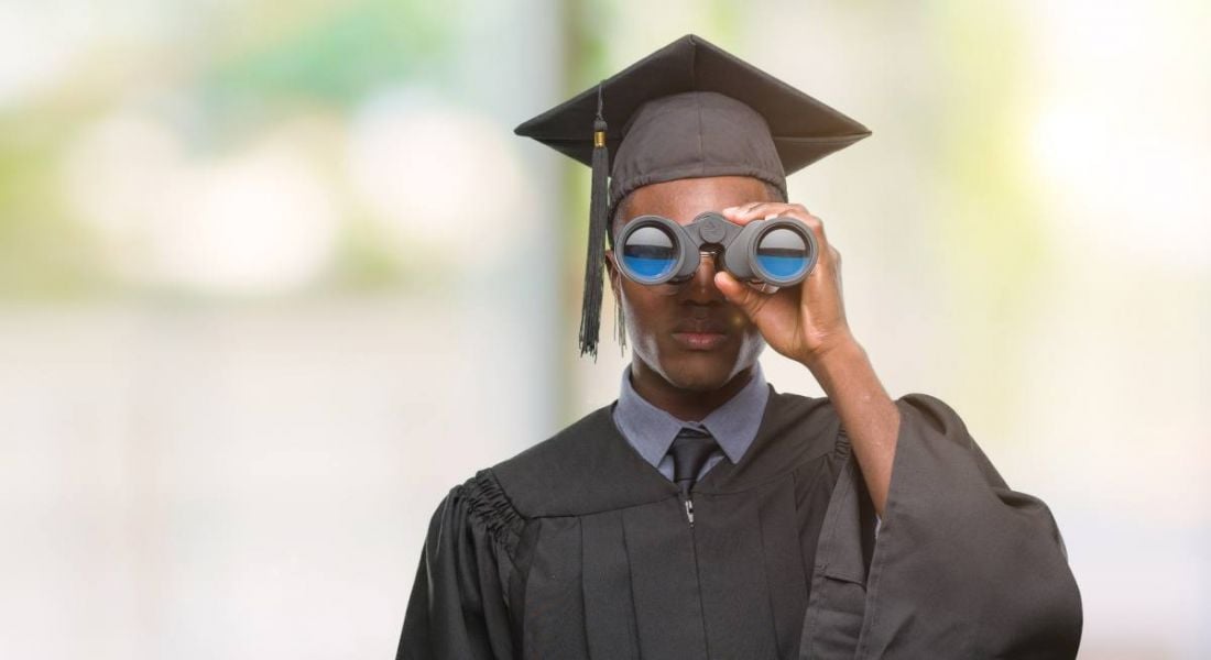 Young male student holding binoculars looking for best employers. He is wearing a graduate's robe and cap.