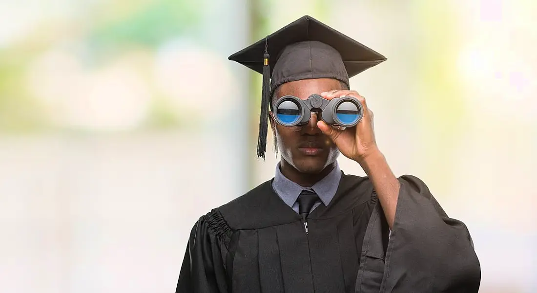 Young male student holding binoculars looking for best employers. He is wearing a graduate's robe and cap.