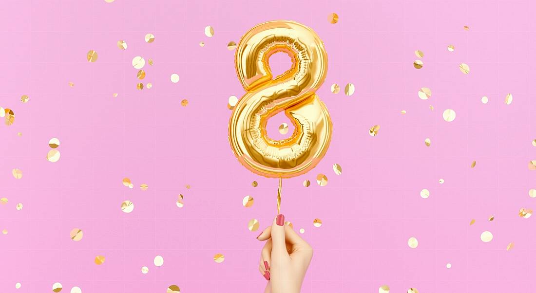 A hand holds up a gold foil balloon shaped like the number eight against a pink backdrop speckled with gold confetti.