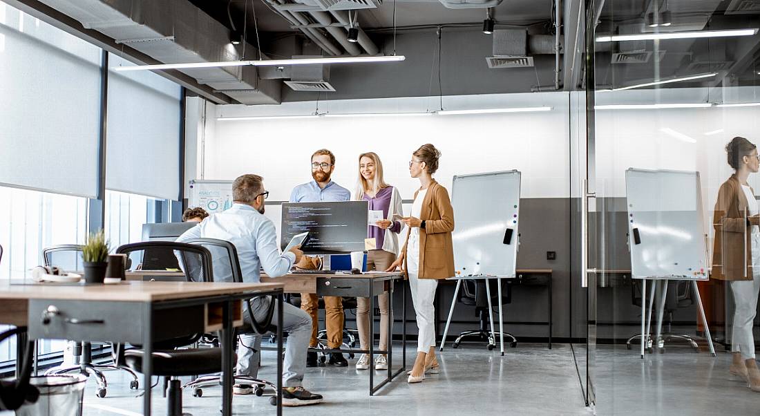 A group of people gathered standing talking around desks in a bright, modern office.