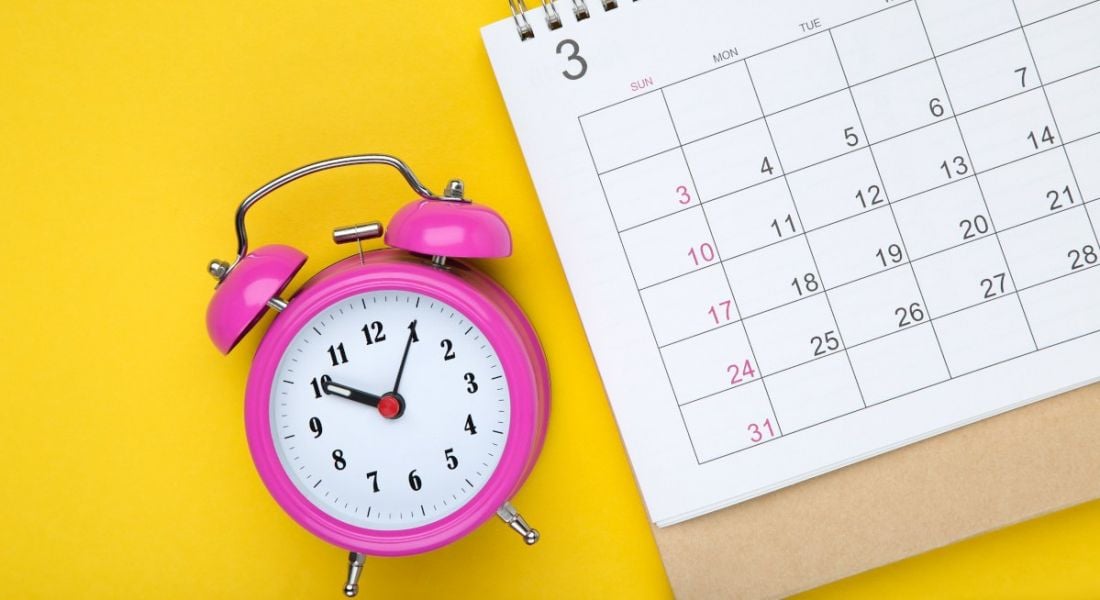 A pink clock is lying on a yellow background with a calendar page in a meetings schedule concept.