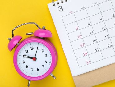 A pink clock is lying on a yellow background with a calendar page in a meetings schedule concept.