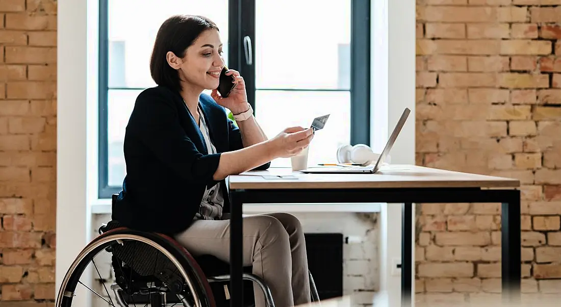 A woman using a wheelchair at a desk in a bright airy room. She is on the phone and has a laptop on the desk.