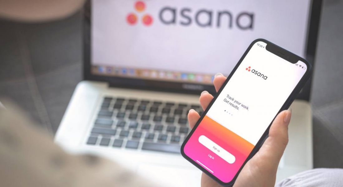 Woman using the Asana app on her smartphone while she has her laptop in front of her with Asana's logo on the screen.