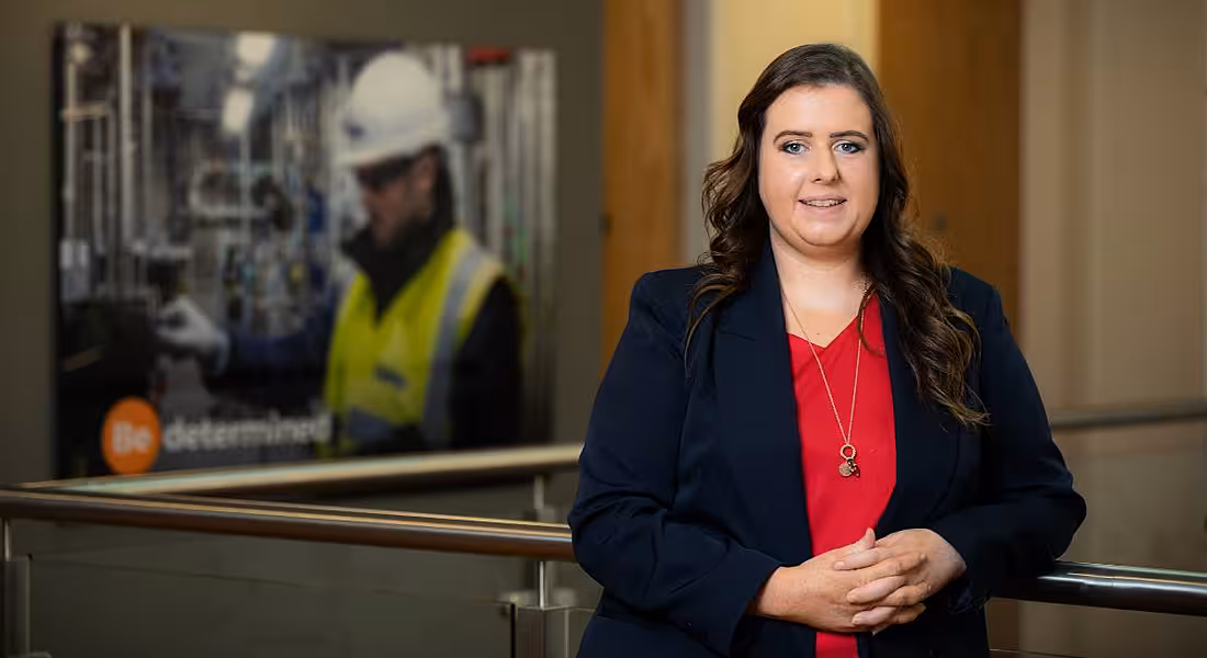 A woman stands in front of a large photograph of a man working as an electrical engineer, wearing a hard hat and high-vis vest.