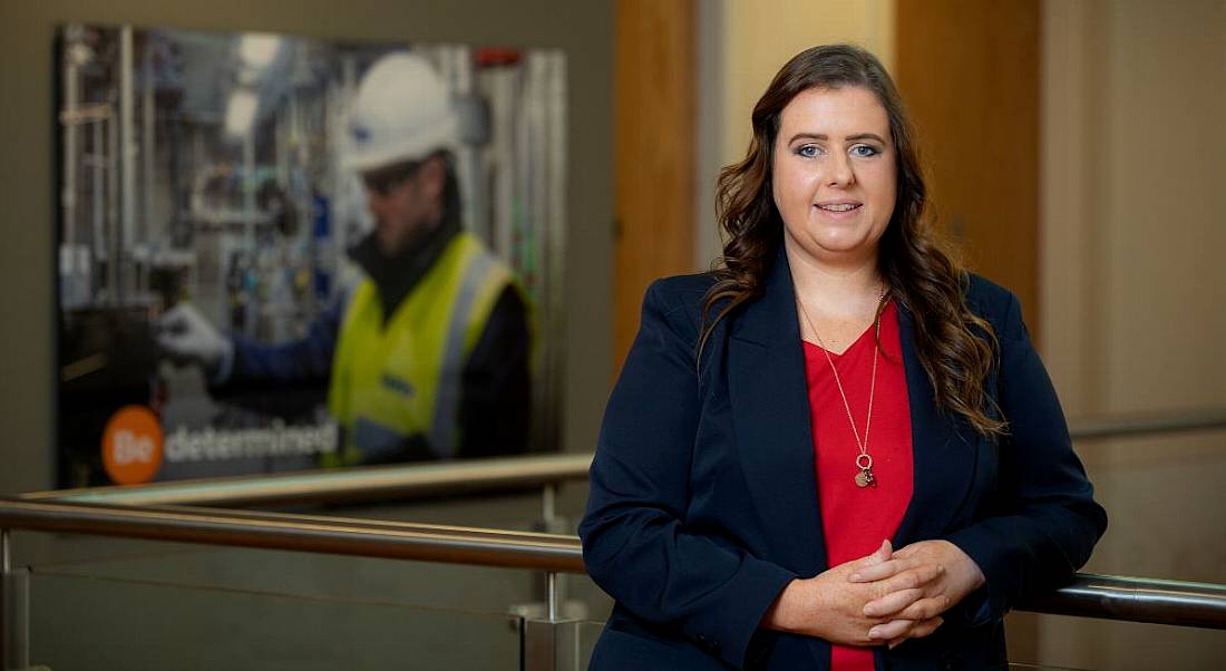 A woman stands in front of a large photograph of a man working as an electrical engineer, wearing a hard hat and high-vis vest.