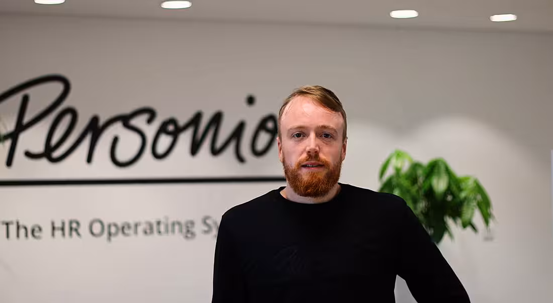 A young man smiles at the camera in an office. The wall behind him has the Personio logo on it.