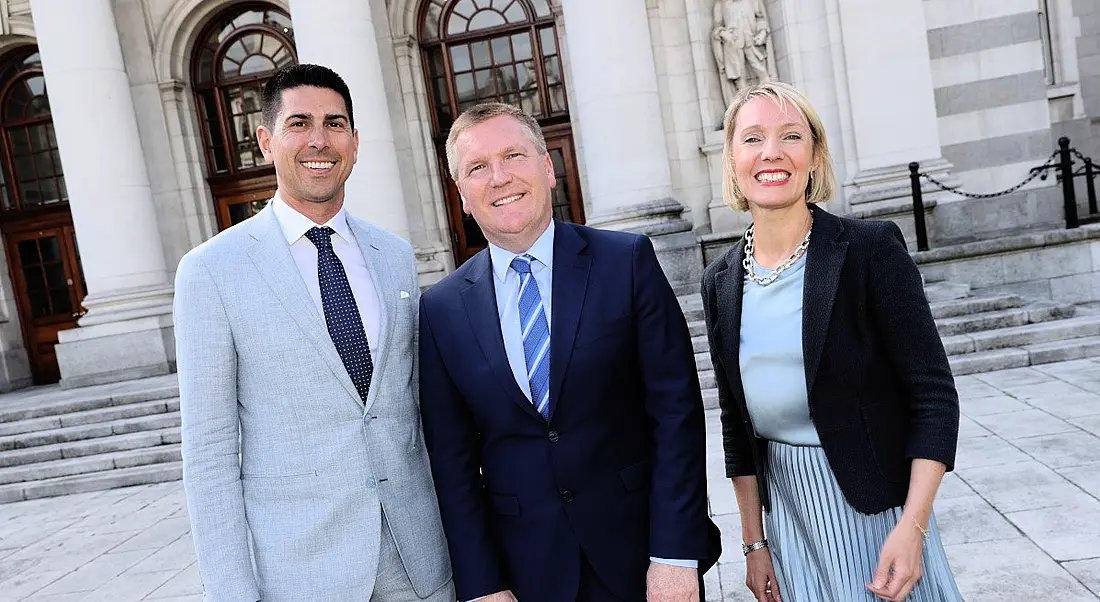 Two men and a woman standing in formal clothing with a grey building behind them.