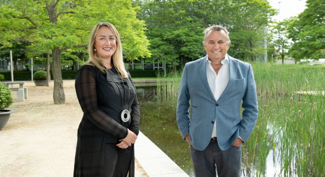 Olivia Bushe, CEO of FlowForma, and John Purdy, non-executive director of FlowForma, stand outside by a pond with green trees in the background.