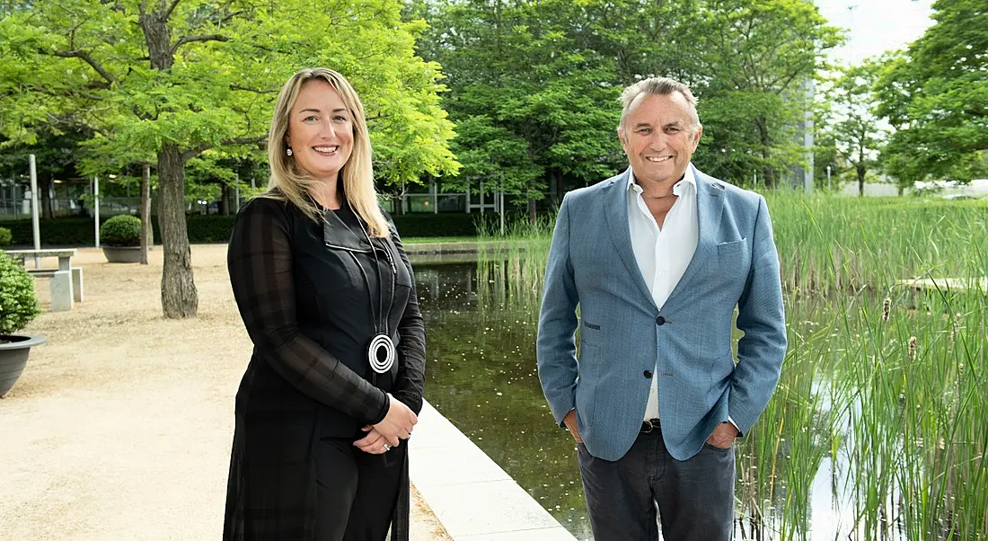 Olivia Bushe, CEO of FlowForma, and John Purdy, non-executive director of FlowForma, stand outside by a pond with green trees in the background.