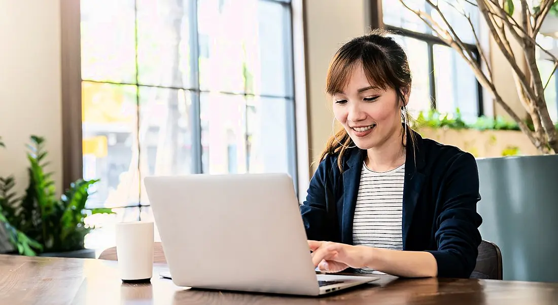 A woman working at a laptop in a brightly lit room smiling as she types.