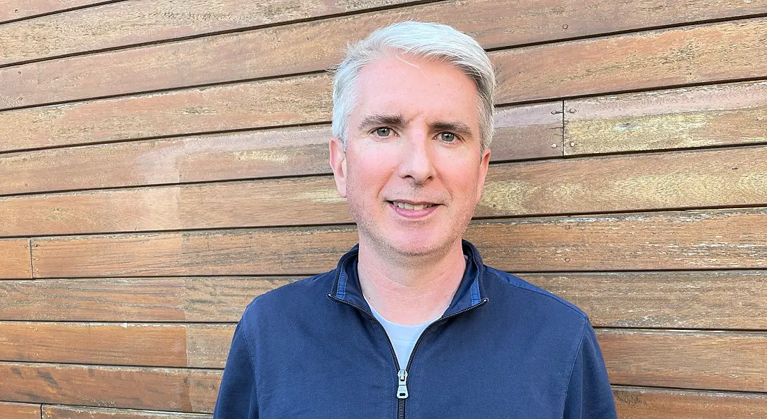 A man in a blue shirt smiles at the camera while standing in front of a brown wooden wall. He is John Walsh of Intel.