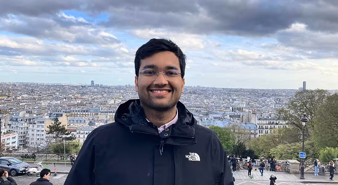 A young man wearing a navy hoody smiles at the camera against the backdrop of a city. He is a product engineer at Intercom.