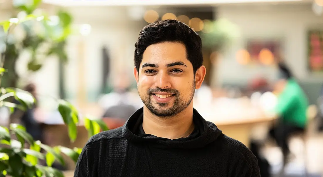 A young man wearing a black jumper smiling at the camera. Behind him is a plant in a bright, out-of-focus office space.