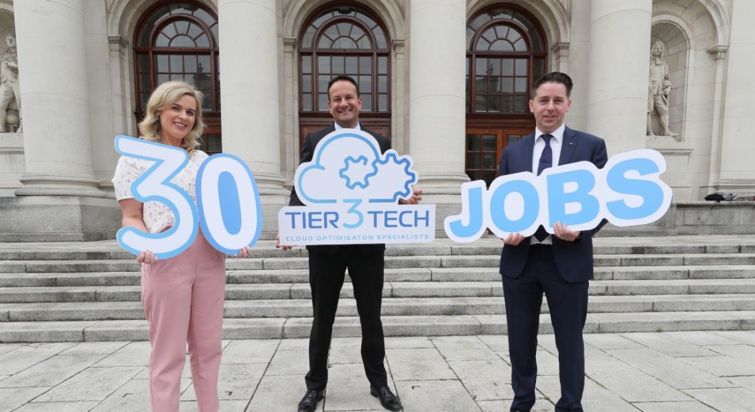 A woman and two men standing outside a large beige stone building holding signs announcing Tier3Tech is hiring.
