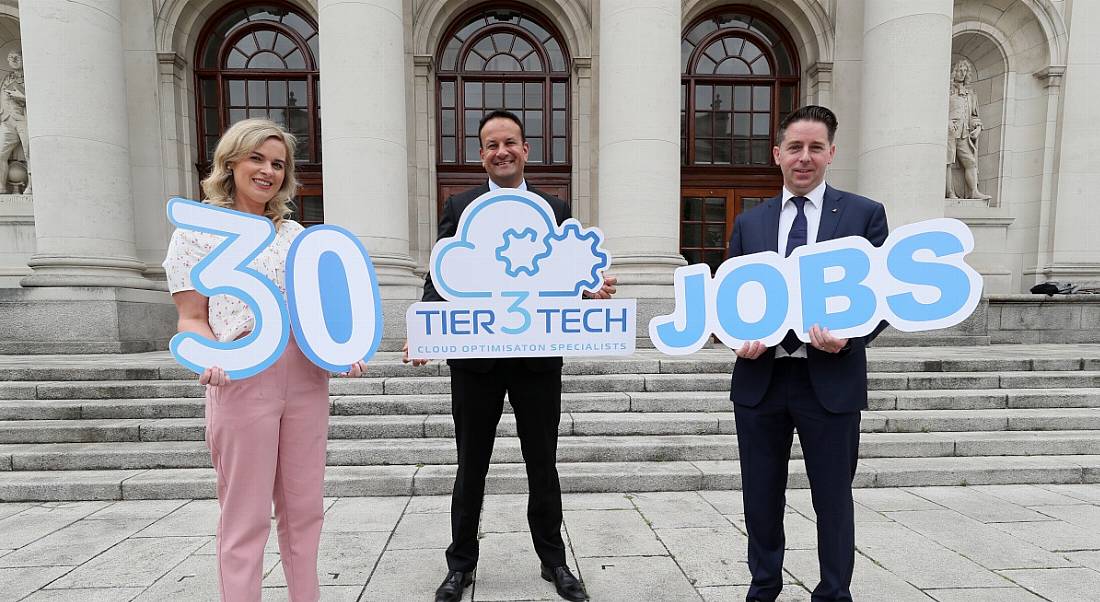 A woman and two men standing outside a large beige stone building holding signs announcing Tier3Tech is hiring.