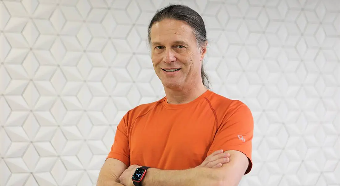 Yahoo's Nate McCoy stands against a white wall with a geometric pattern smiling at the camera. He is wearing an orange t-shirt.