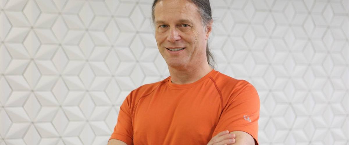 Yahoo's Nate McCoy stands against a white wall with a geometric pattern smiling at the camera. He is wearing an orange t-shirt.
