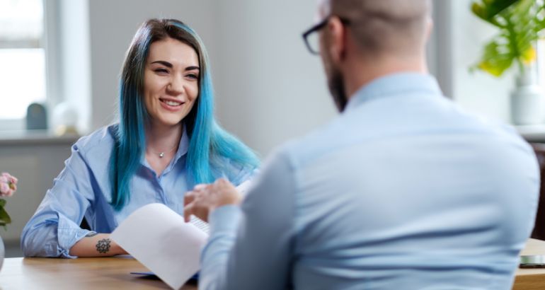 A young woman with dyed blue smiling in a job interview while a man looks through her CV.
