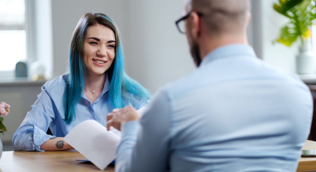 A young woman with dyed blue smiling in a job interview while a man looks through her CV.