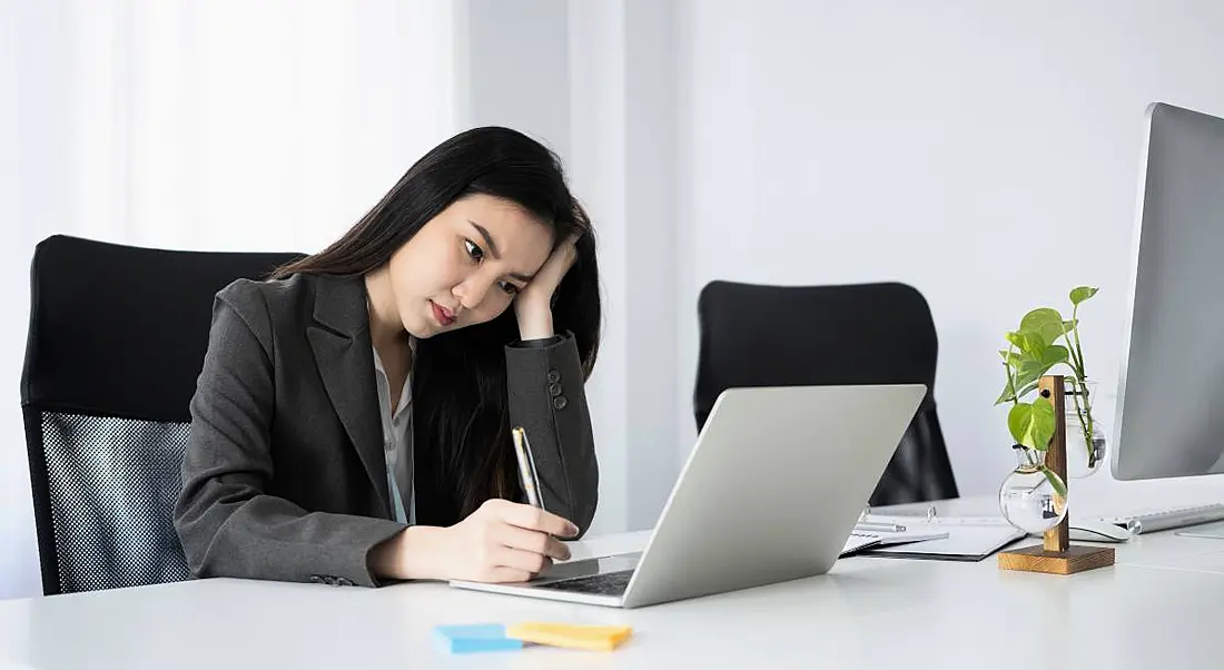 A stressed out young woman sitting in front of a laptop and taking notes.