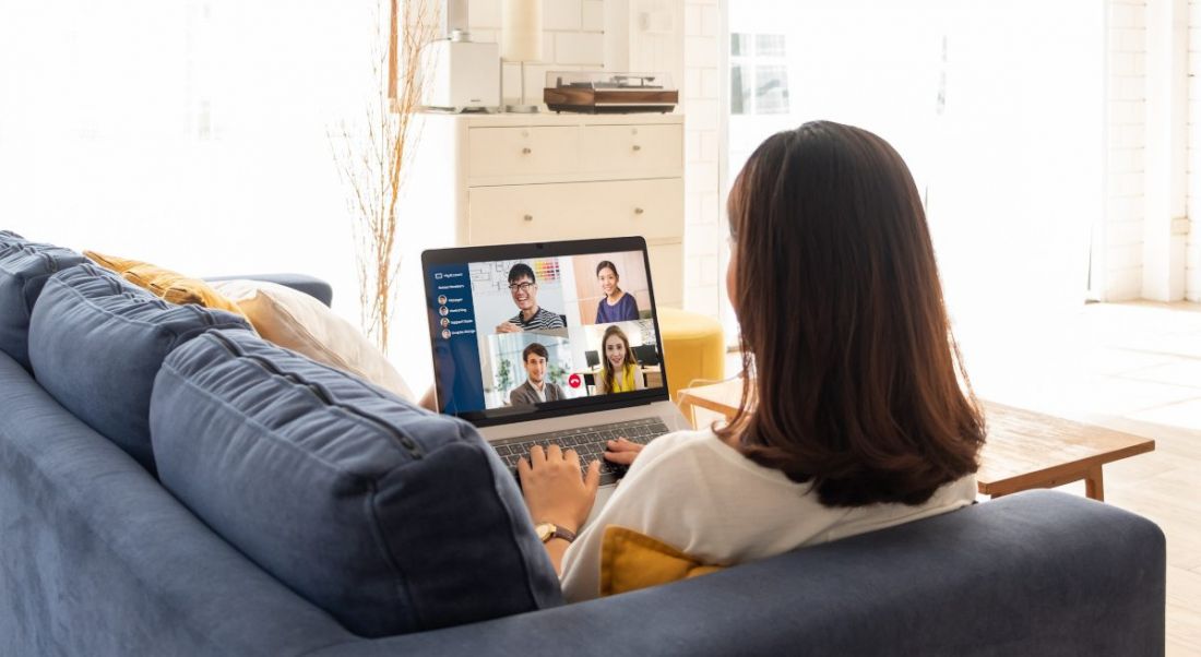 A woman sitting on a couch in a brightly lit living room. She’s working on a laptop that shows four people on a conference call, depicting a remote-first culture.