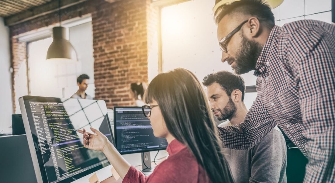 A woman software developer points to a screen of code while two men software developers look on.