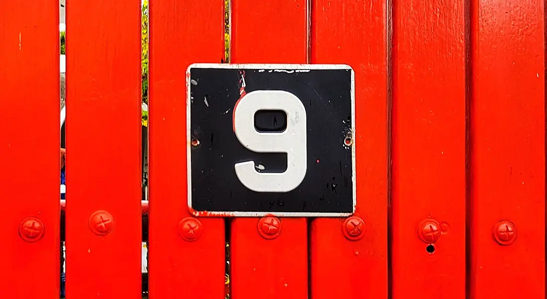 A black sign with a white number nine on it. The sign is on a bright red wooden fence.