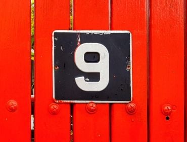 A black sign with a white number nine on it. The sign is on a bright red wooden fence.