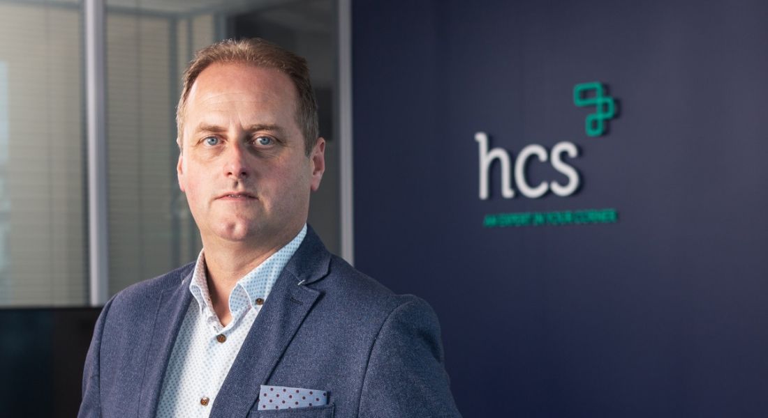 Neil Phelan dressed in a navy suit with spotted shirt and pocket square stands in front of a wall branded with the HCS logo.