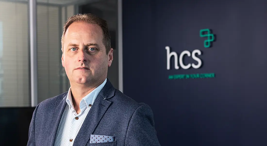 Neil Phelan dressed in a navy suit with spotted shirt and pocket square stands in front of a wall branded with the HCS logo.