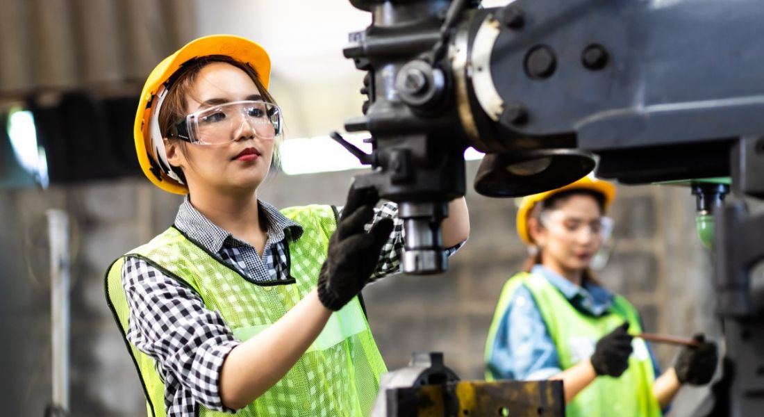 Two women working in manufacturing with machines wearing safety goggles and hard hats.
