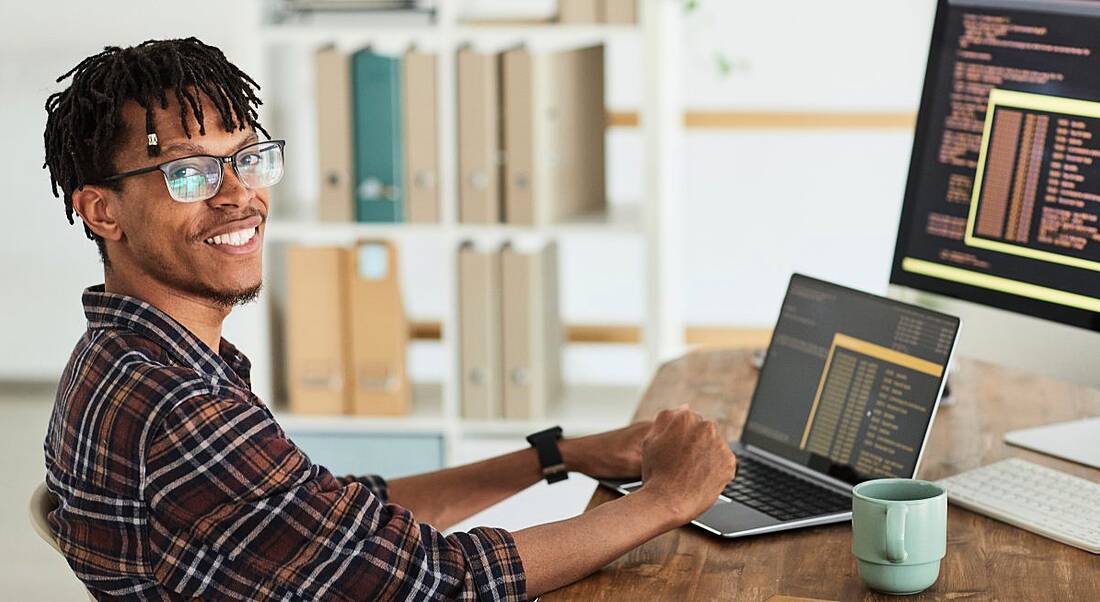 Young man sitting at a desk typing on a laptop with a mug beside him and a desktop computer behind the laptop.