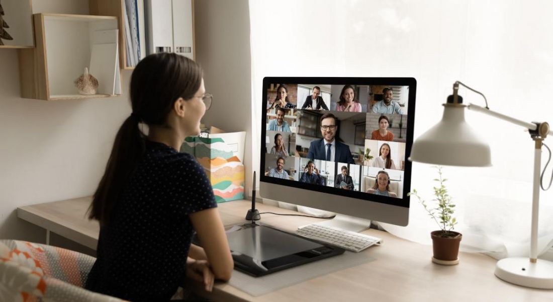 A woman in front of a computer screen with other people on a video call. The computer is on a wooden desk with a lamp next to it, in a home room environment, used to show the concept of remote working.