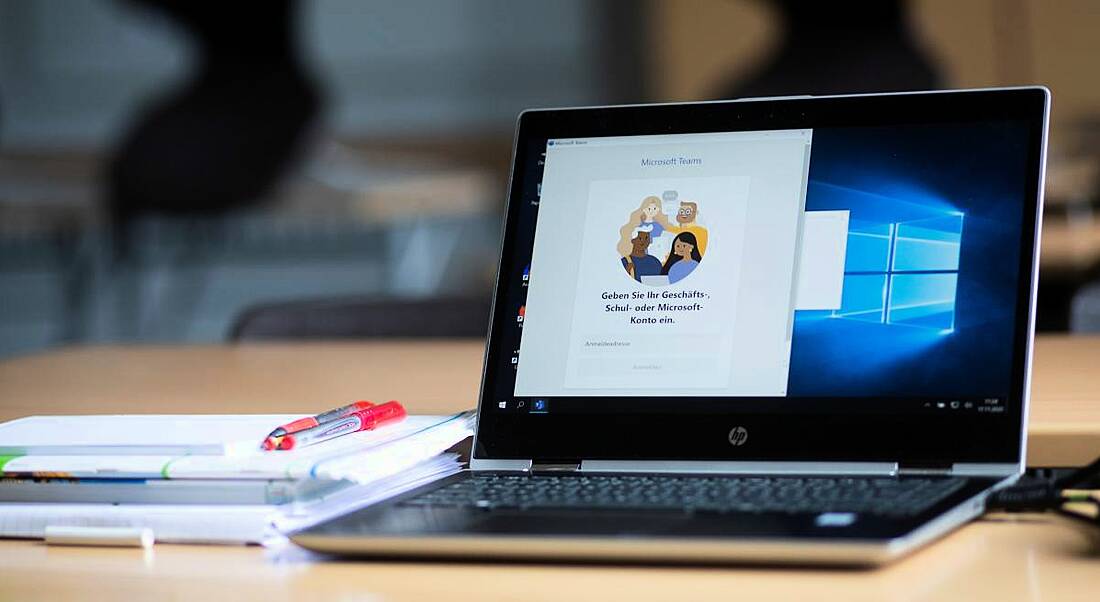 Laptop with Microsoft Teams open on its screen and a notebook and pen beside it on a table.