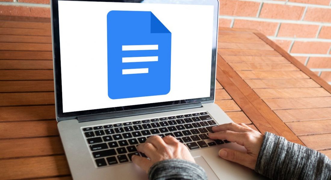 A person typing on a laptop with the Google Docs logo displayed on the screen. There is a red brick wall in the background.