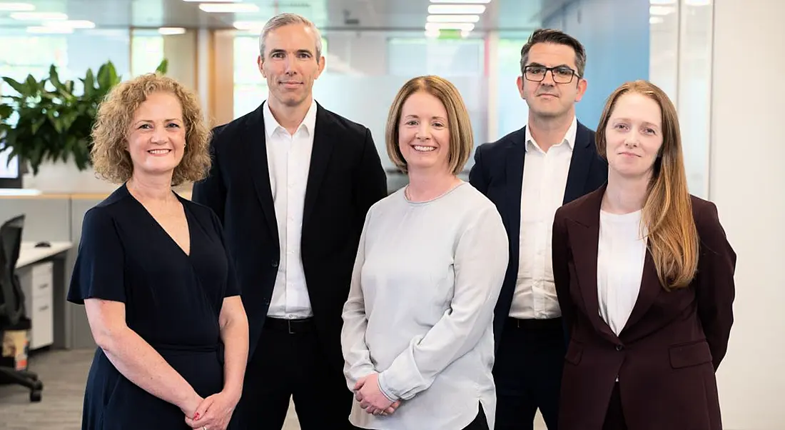 Five people who work at BearingPoint consultancy company standing in a group in a brightly lit office.