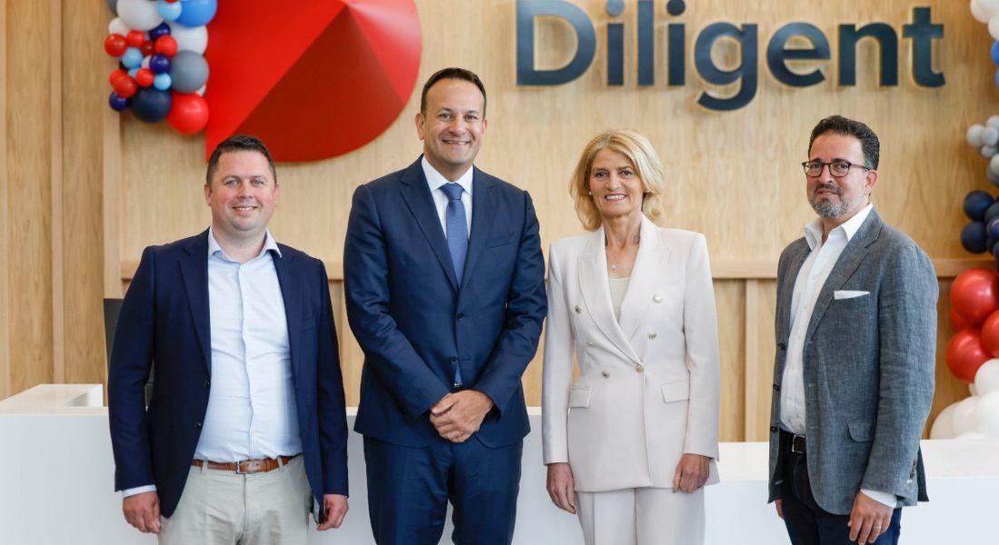 Four people, three men and one woman, at the opening of SaaS company Diligent and its Galway office. They are standing against a wooden panelled wall with balloons in the background.