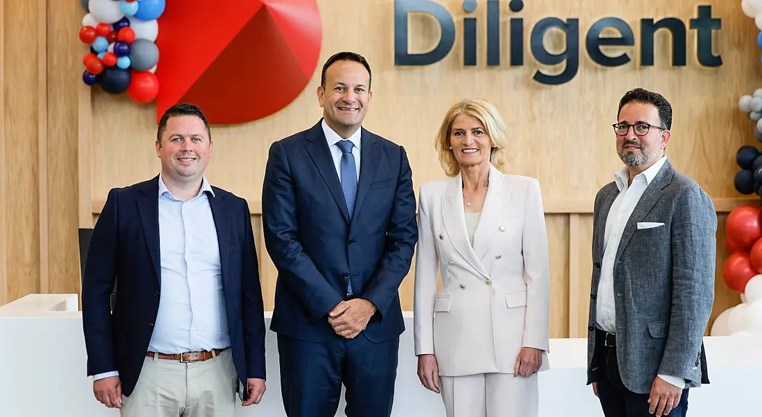 Four people, three men and one woman, at the opening of SaaS company Diligent and its Galway office. They are standing against a wooden panelled wall with balloons in the background.