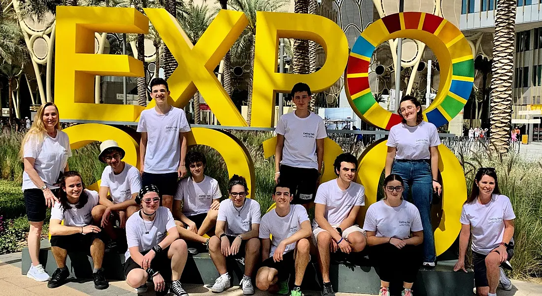 A large group of students in white T-shirts stand in front of large yellow sign that says ‘Expo’.