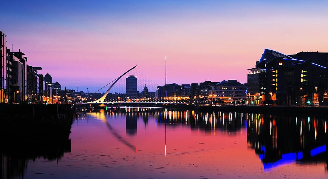 Dublin City Centre skyline with Samuel Beckett bridge at dusk. The sky goes from blue to pink and is reflected in the Liffey, while buildings are lit up.