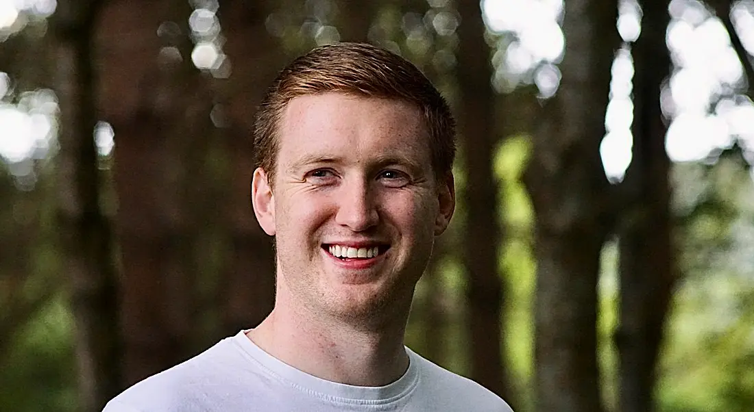 A young man wearing a white t-shirt smiling at the camera. There are many trees behind him.